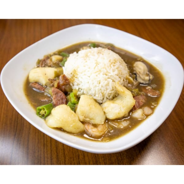 A white bowl of seafood gumbo with rice, shrimp, sausage, and okra on a wooden table.
